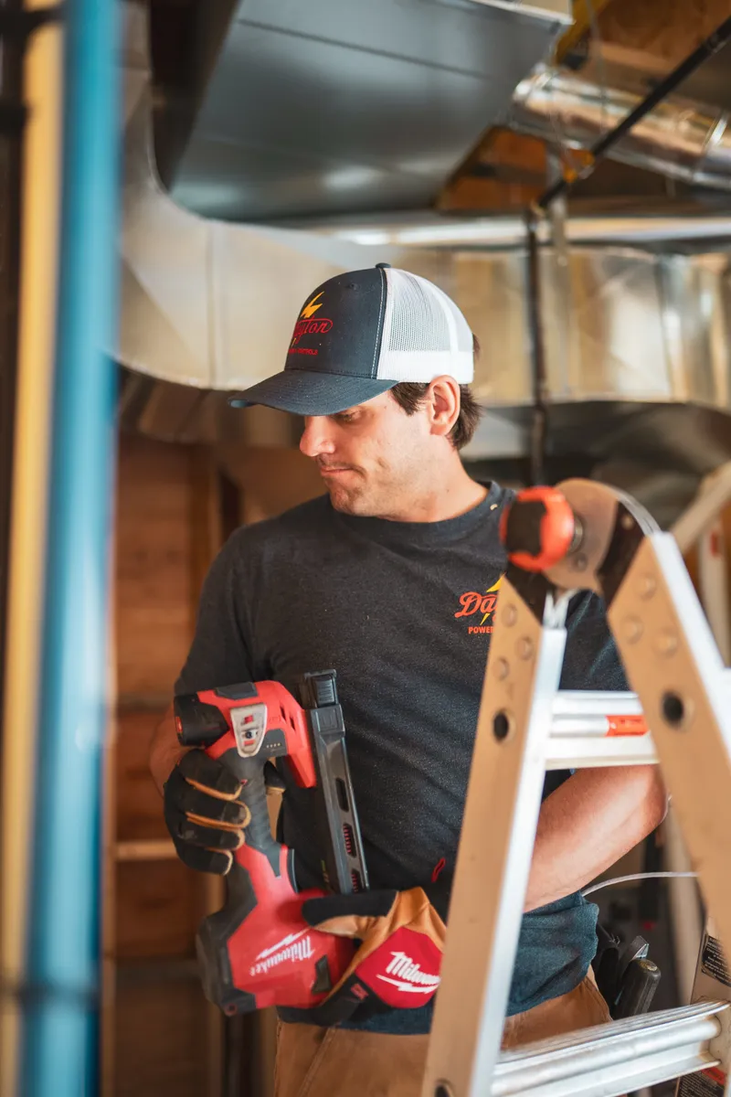 Dayton Power electrician in a branded hat working from a ladder beneath metal ductwork with a cordless stapler